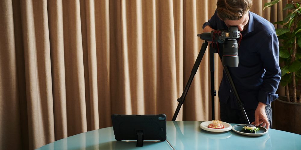 A photoshoot of food plates being captured by a standing man holding the camera and wirelessly tethering to tablet that stands next to the plates
